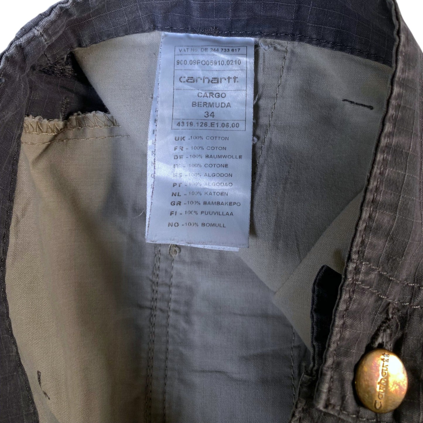 Close-up of the inside waistband of a Carhartt cargo jacket in Carhartt Bermuda twill, showing the white brand label, interior lining, and a brass button.