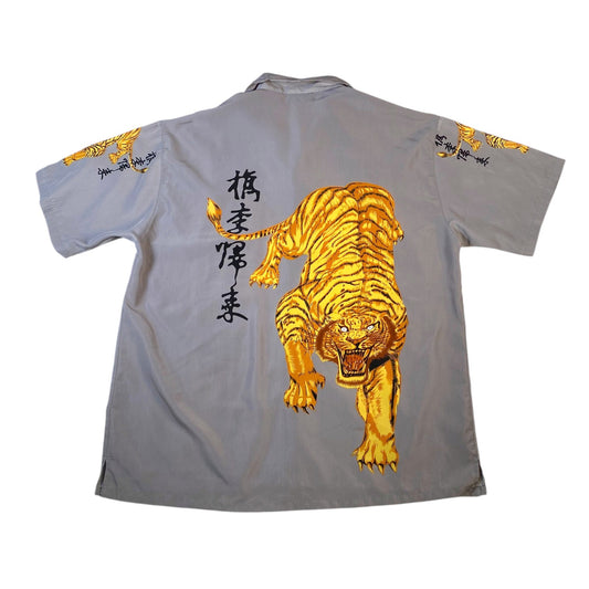 A light gray short-sleeve button-up shirt shown flat against a white background, featuring a large golden tiger graphic on the back, black Japanese characters, and smaller tiger motifs on both sleeves.