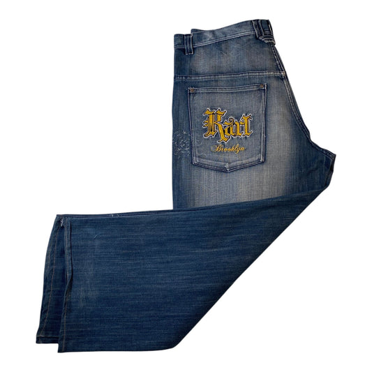 A pair of folded dark blue denim jeans on a plain white background, with the back pocket of one leg showing a yellow embroidered logo that reads 'Kat Brooklyn'.