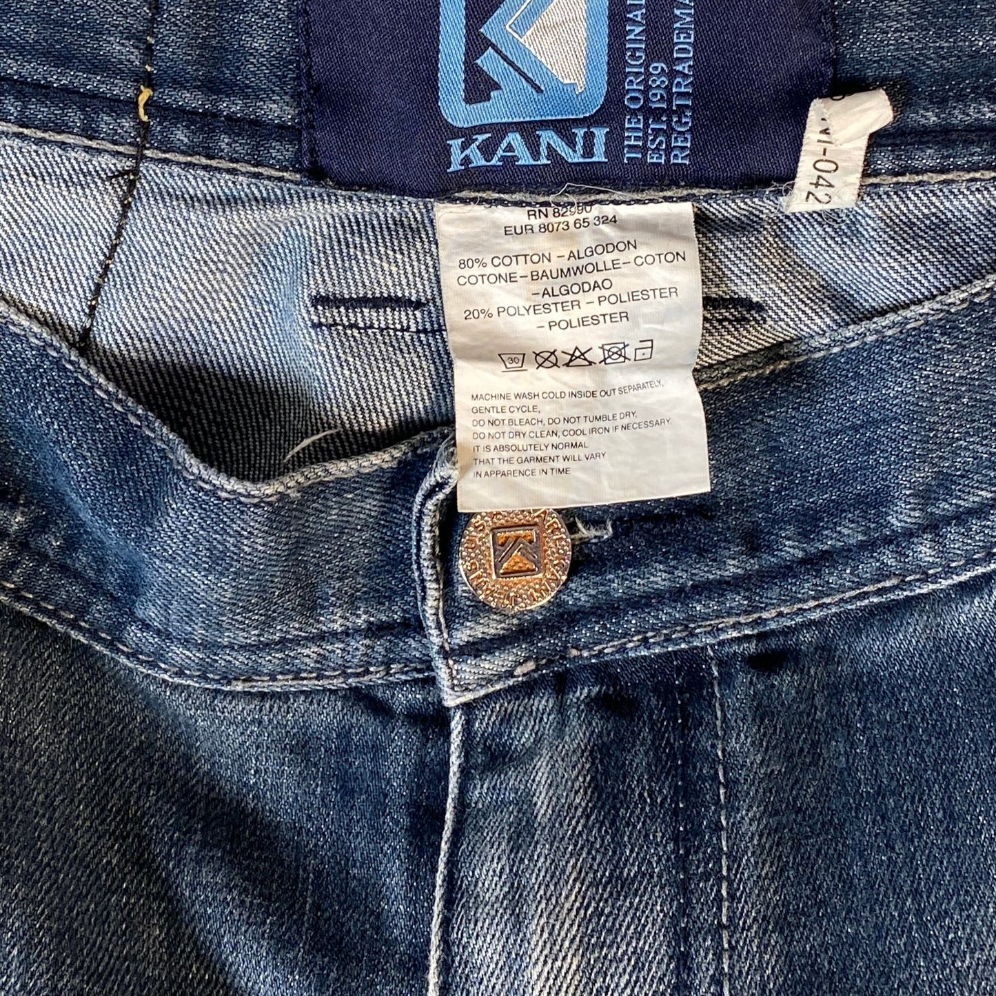 Close-up of blue denim jeans showing the inner waistband, Kani brand label, care instructions tag, and a brass button.