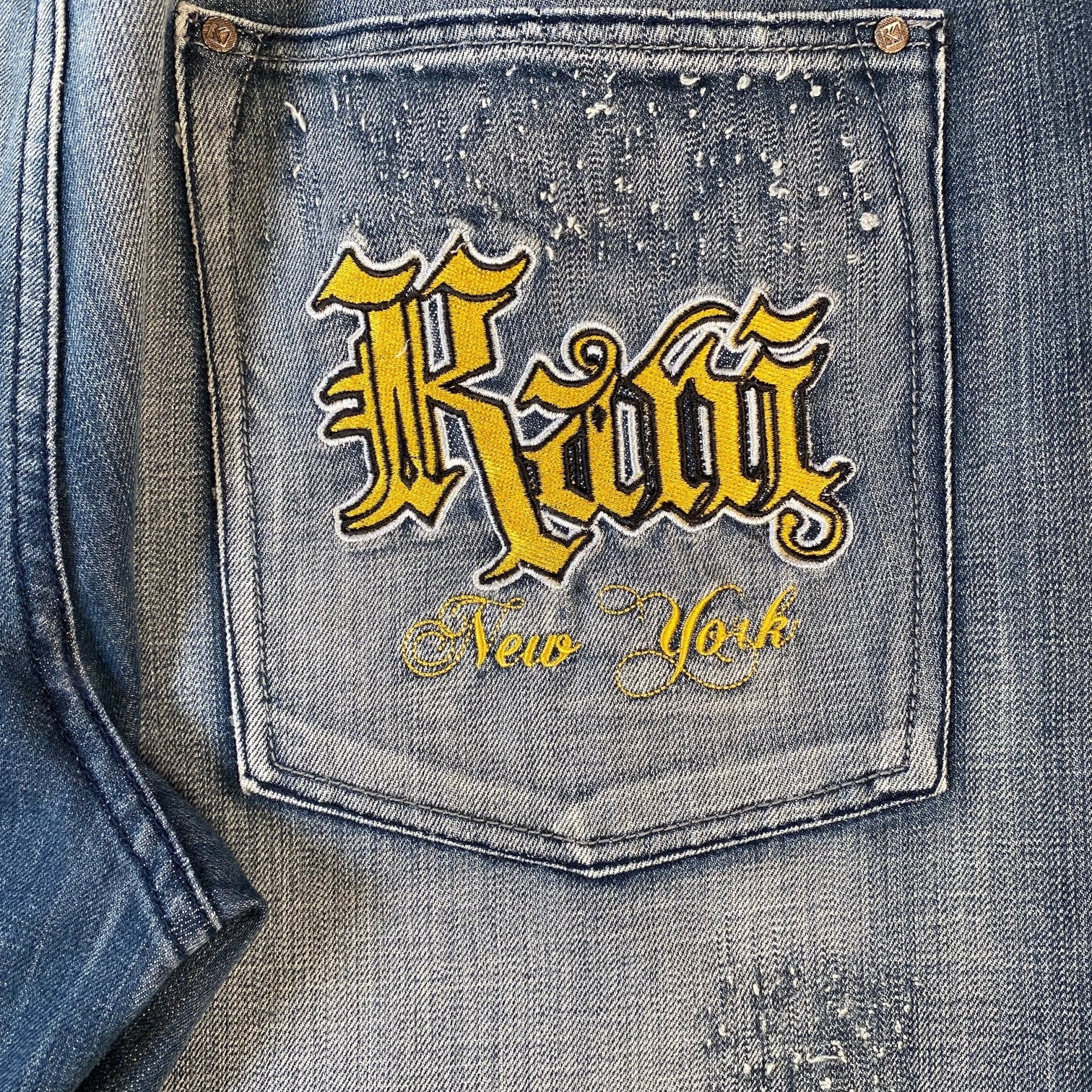 Close-up of a distressed blue denim jacket focusing on the back pocket, featuring a prominent yellow embroidered patch with the gothic-style text 'Kawi' and 'New York' below it.