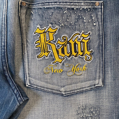 Close-up of a distressed blue denim jacket focusing on the back pocket, featuring a prominent yellow embroidered patch with the gothic-style text 'Kawi' and 'New York' below it.