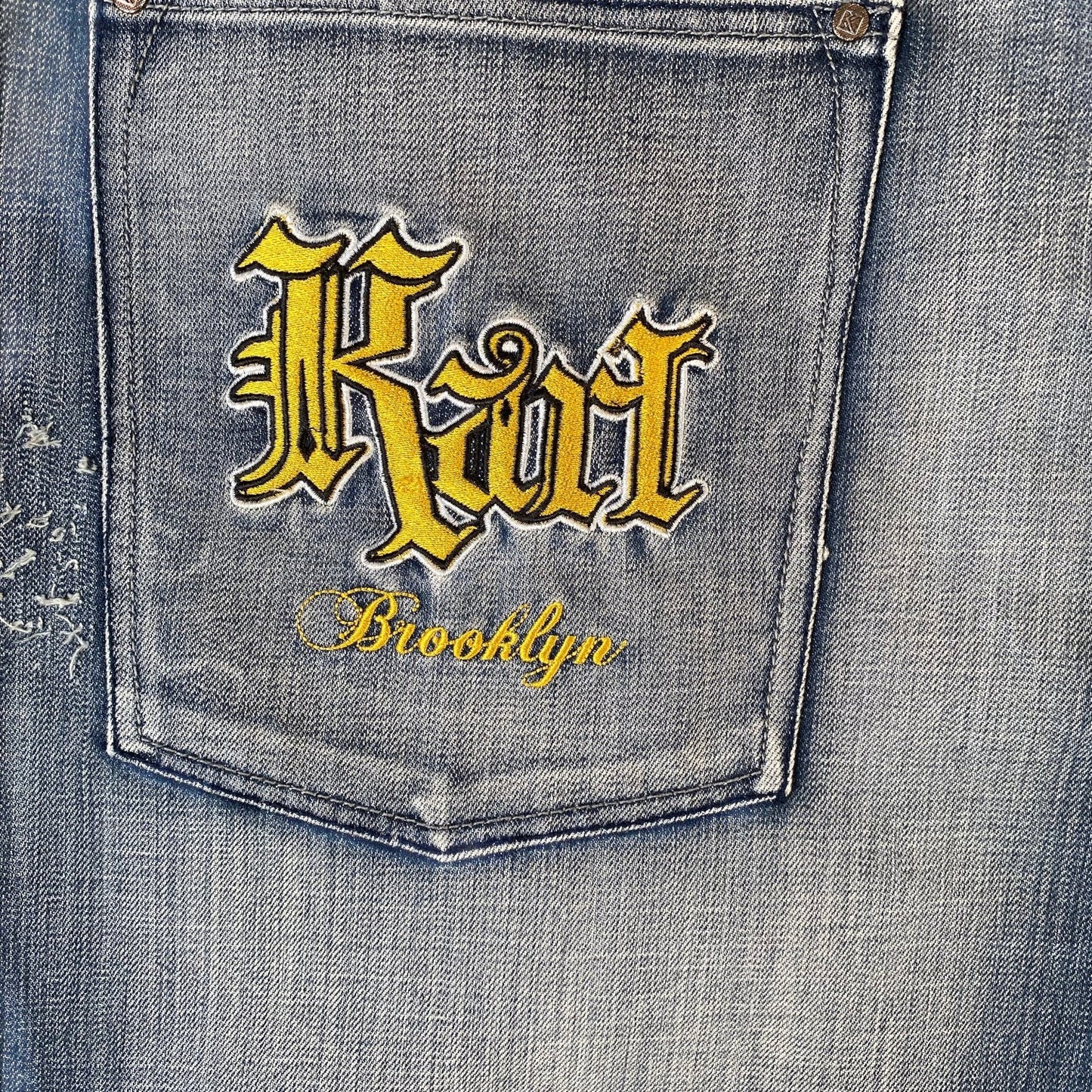 Close-up of a denim jeans back pocket with yellow embroidered gothic-style 'Riot' lettering and 'Brooklyn' script on a faded blue wash.