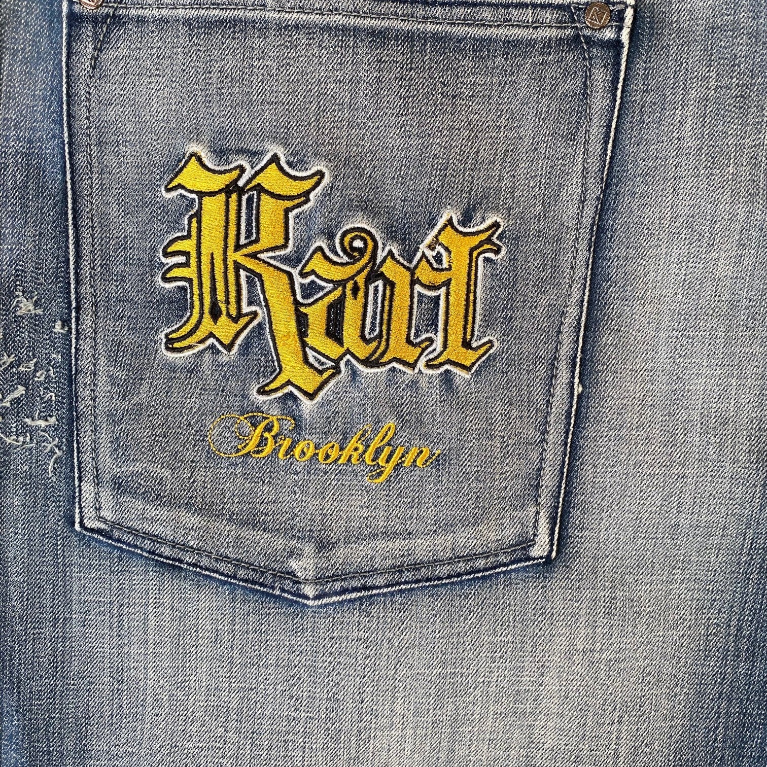 Close-up of a denim jeans back pocket with yellow embroidered gothic-style 'Riot' lettering and 'Brooklyn' script on a faded blue wash.