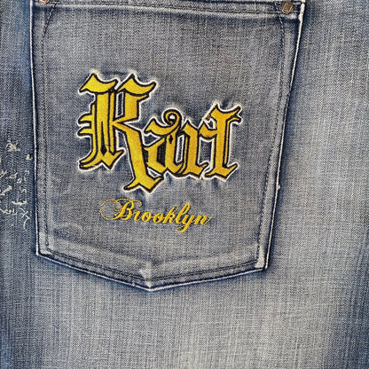 Close-up of a denim jeans back pocket with yellow embroidered gothic-style 'Riot' lettering and 'Brooklyn' script on a faded blue wash.