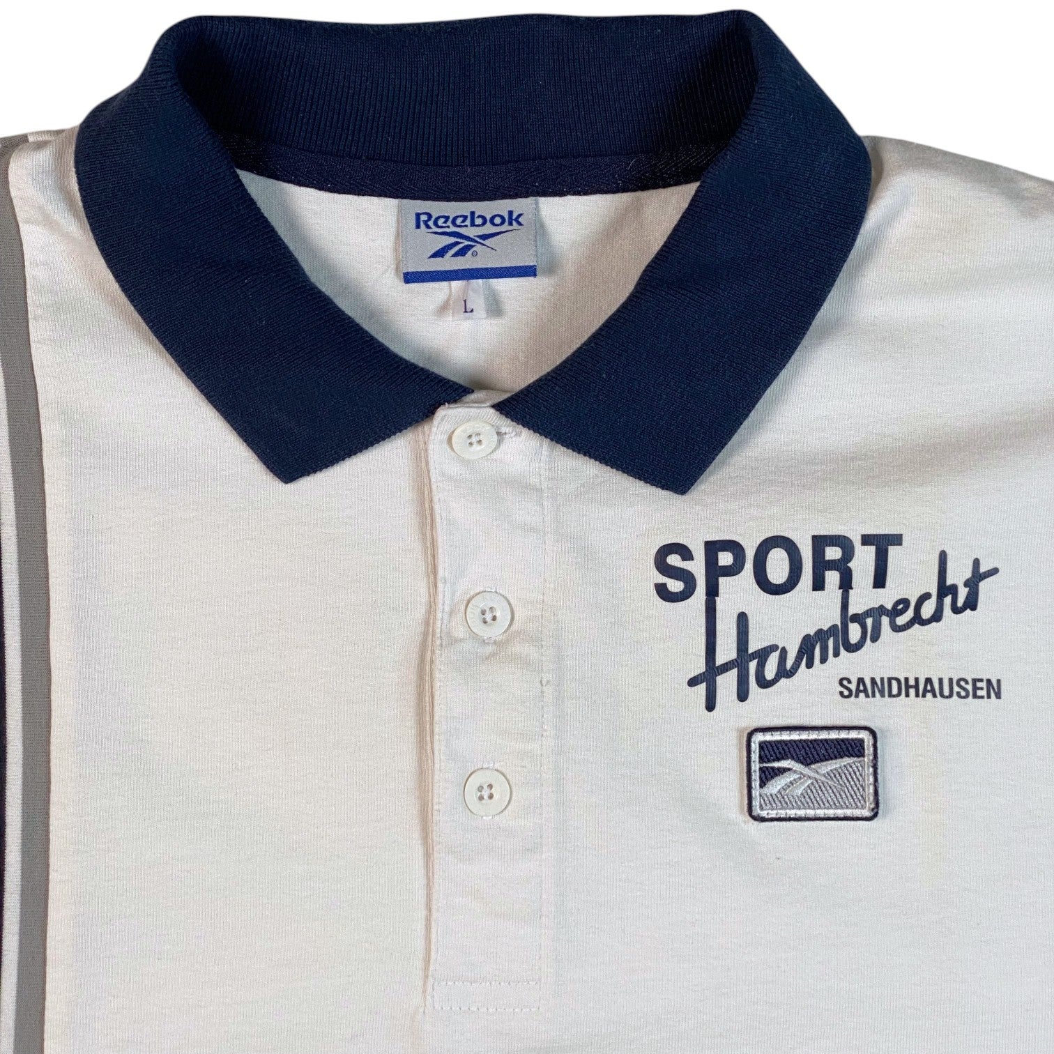 A close-up of a Reebok polo shirt with a white body, navy blue collar, and navy blue printed graphics on the chest reading 'SPORT Hambrecht SANDHAUSEN'.