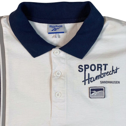 A close-up of a Reebok polo shirt with a white body, navy blue collar, and navy blue printed graphics on the chest reading 'SPORT Hambrecht SANDHAUSEN'.
