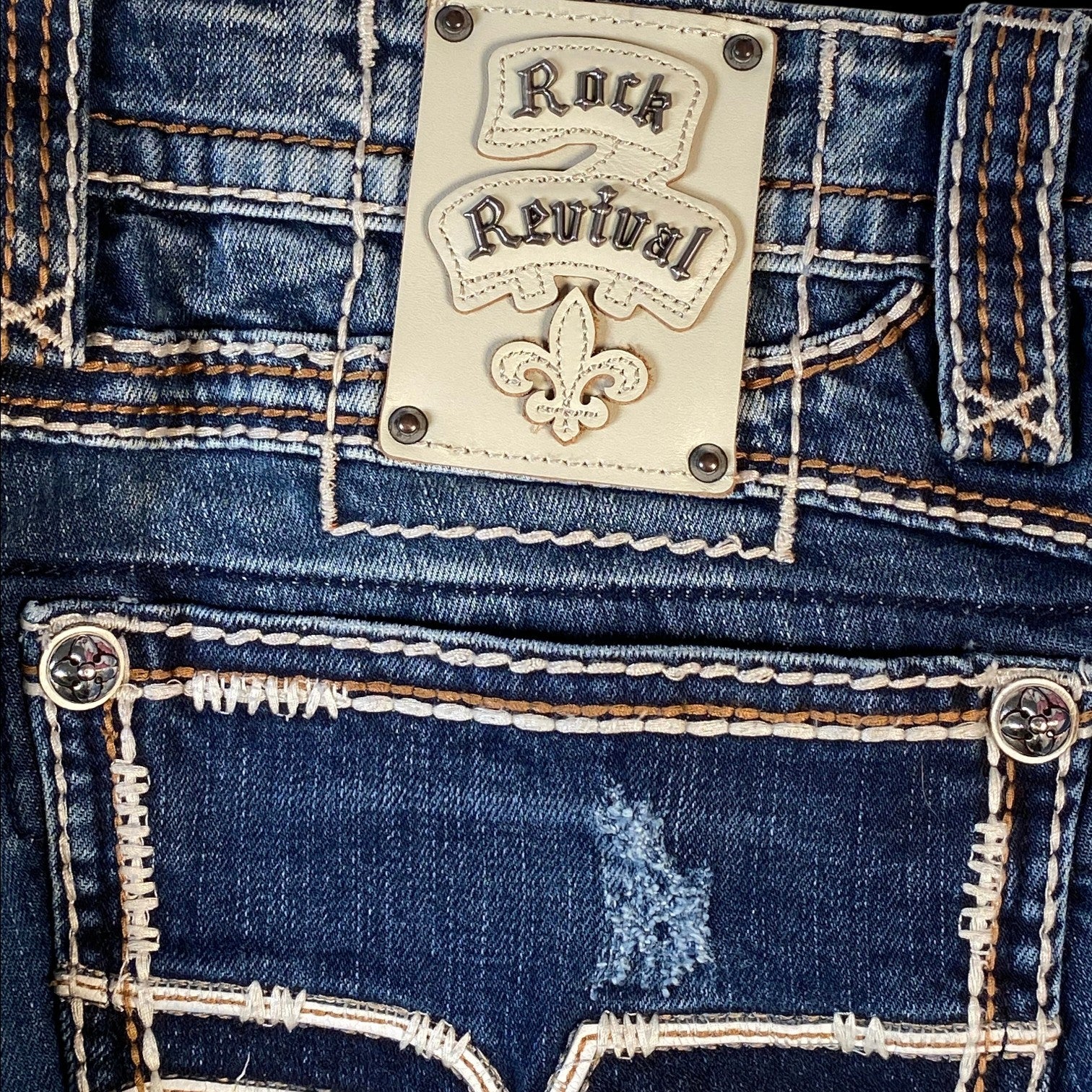Close-up of the back waistband and back pockets of blue denim jeans with contrast stitching, a distressed area on the pocket, and a cream-colored Rock Revival brand patch.