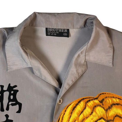 Close-up of a gray short-sleeve button-up shirt with a collar, featuring a black Brother brand label at the inner neck and a yellow and black tiger-stripe graphic across the lower front.