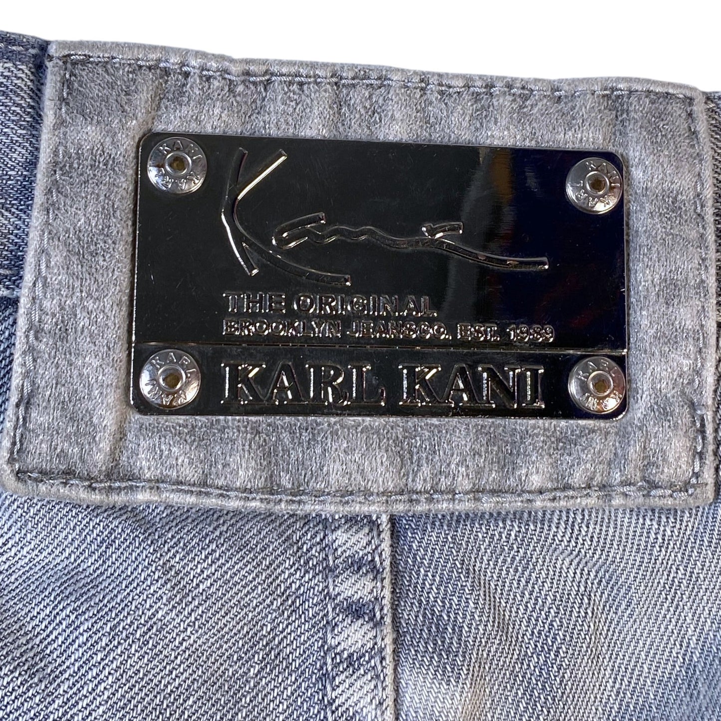 Close-up of a Karl Kani denim product showing a black metal brand plaque with silver rivets on a light blue denim fabric.
