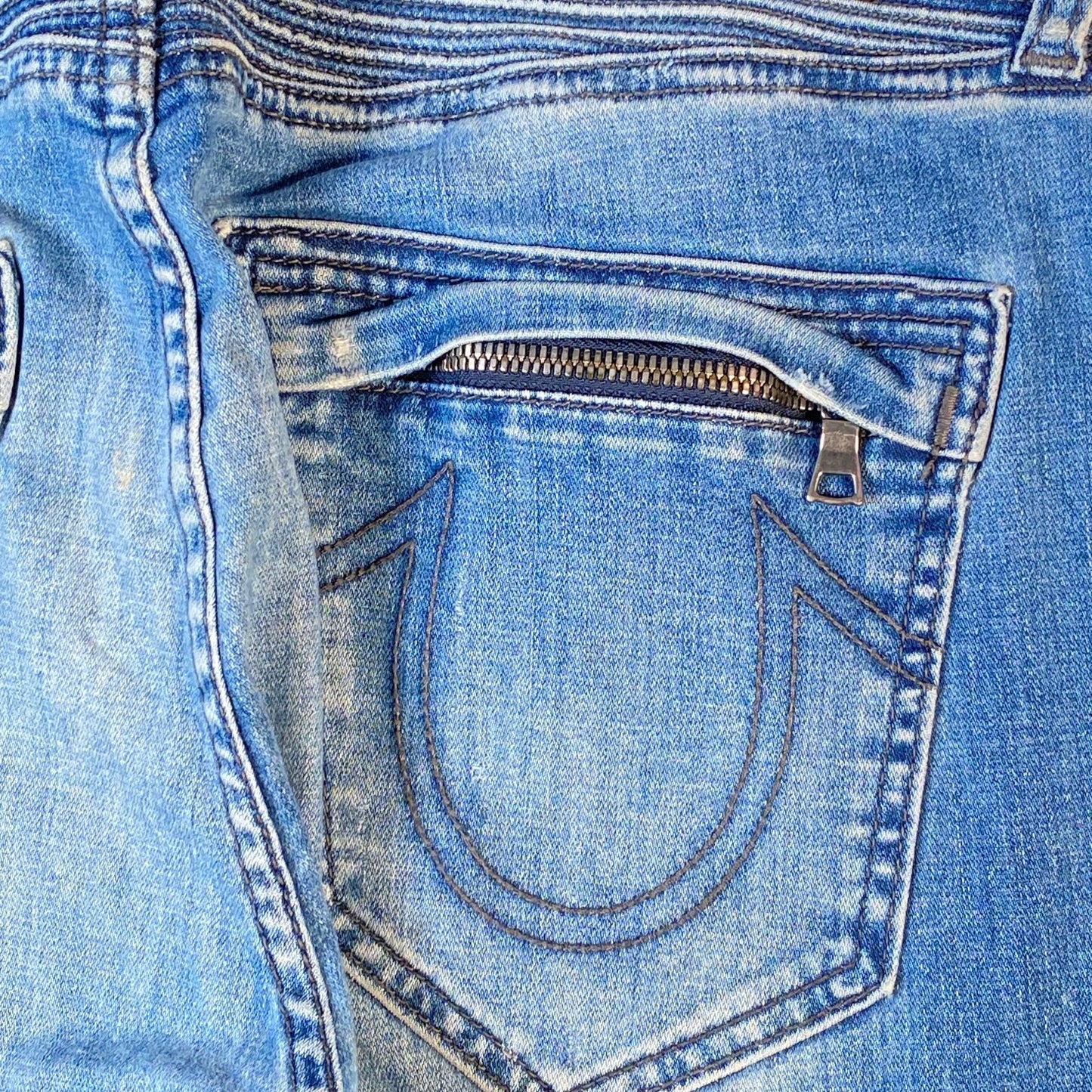 Close-up of the back pocket of light blue denim jeans, showing a zippered pocket with a metal pull tab and contrast stitching.