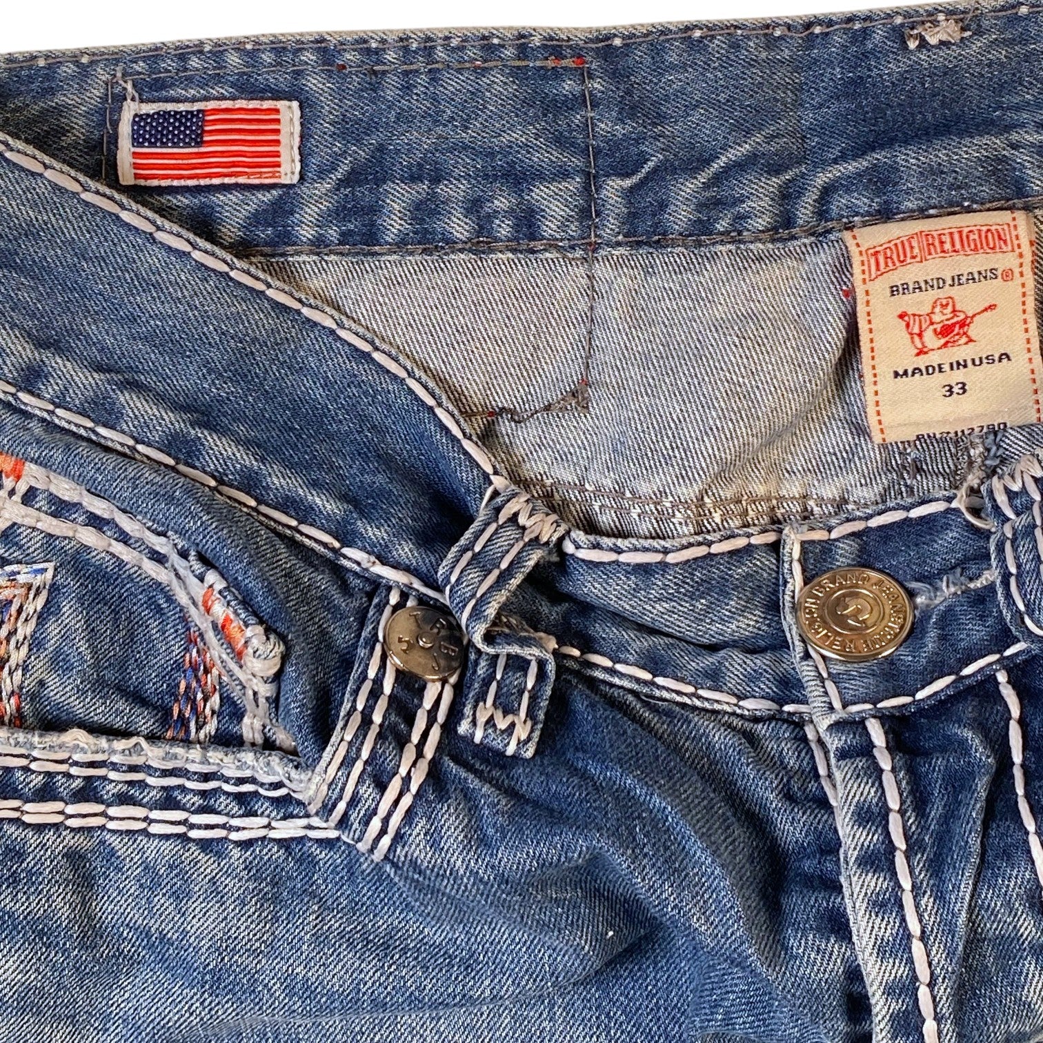 Close-up of the waistband and front details of a pair of blue denim jeans, showing a visible True Religion brand patch, an American flag patch, and contrast stitching.