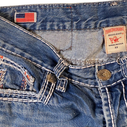 Close-up of the waistband and front details of a pair of blue denim jeans, showing a visible True Religion brand patch, an American flag patch, and contrast stitching.