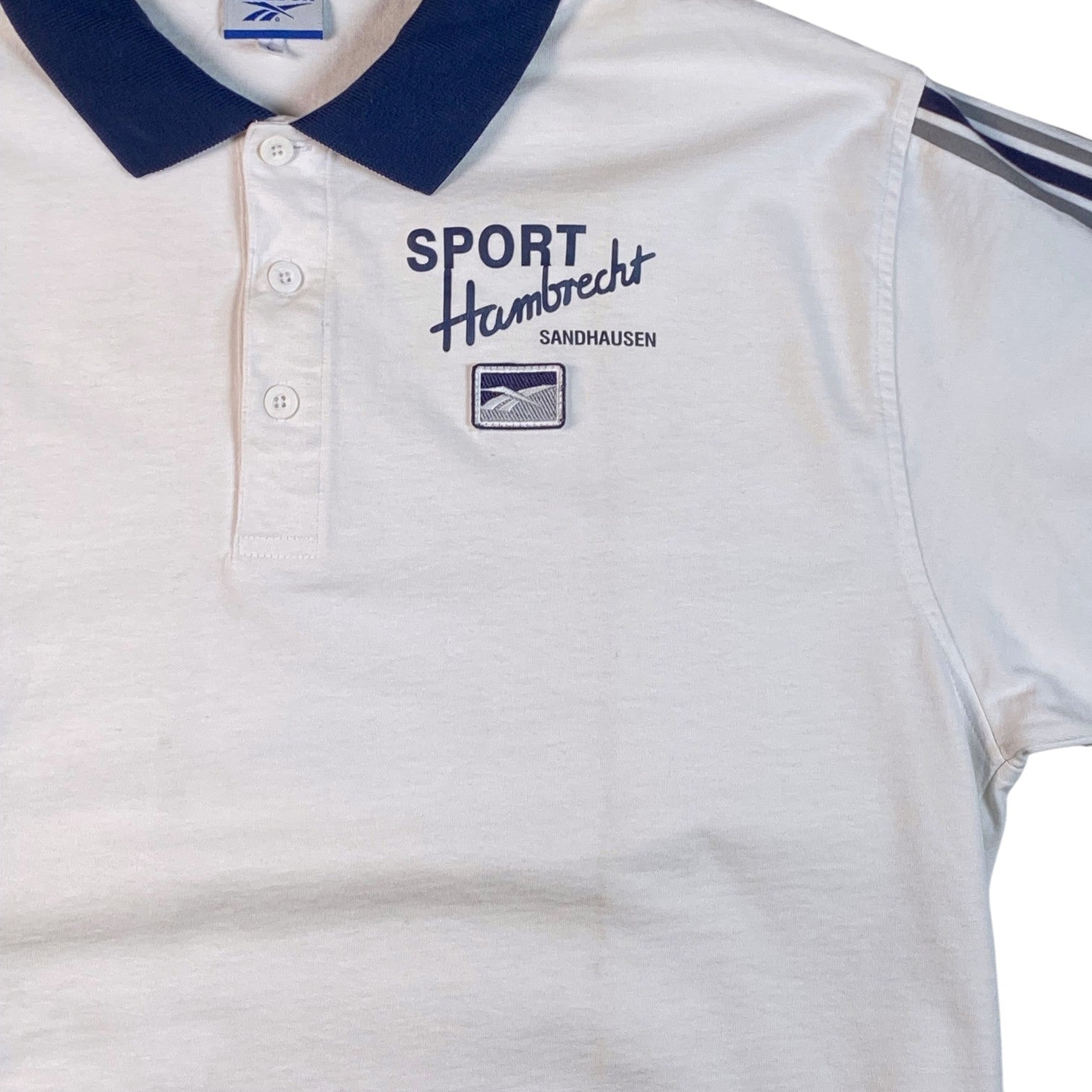 A white short-sleeve polo shirt with a navy blue collar, featuring 'SPORT Hambrecht SANDHAUSEN' and a logo patch on the left chest, and navy stripes on the sleeve.