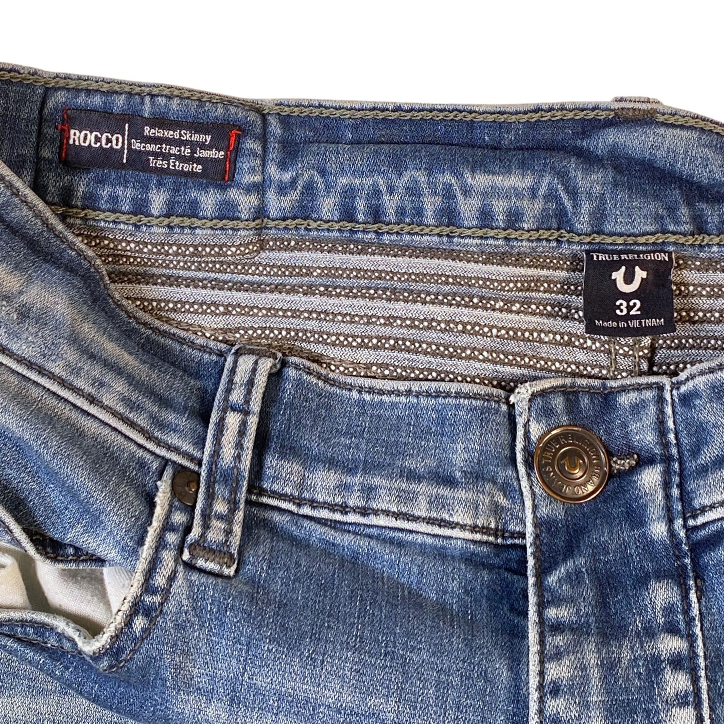 Close-up of the waistband and front closure of blue denim jeans, showing a metal button, belt loops, and two visible brand labels: Rocco and Trtl.