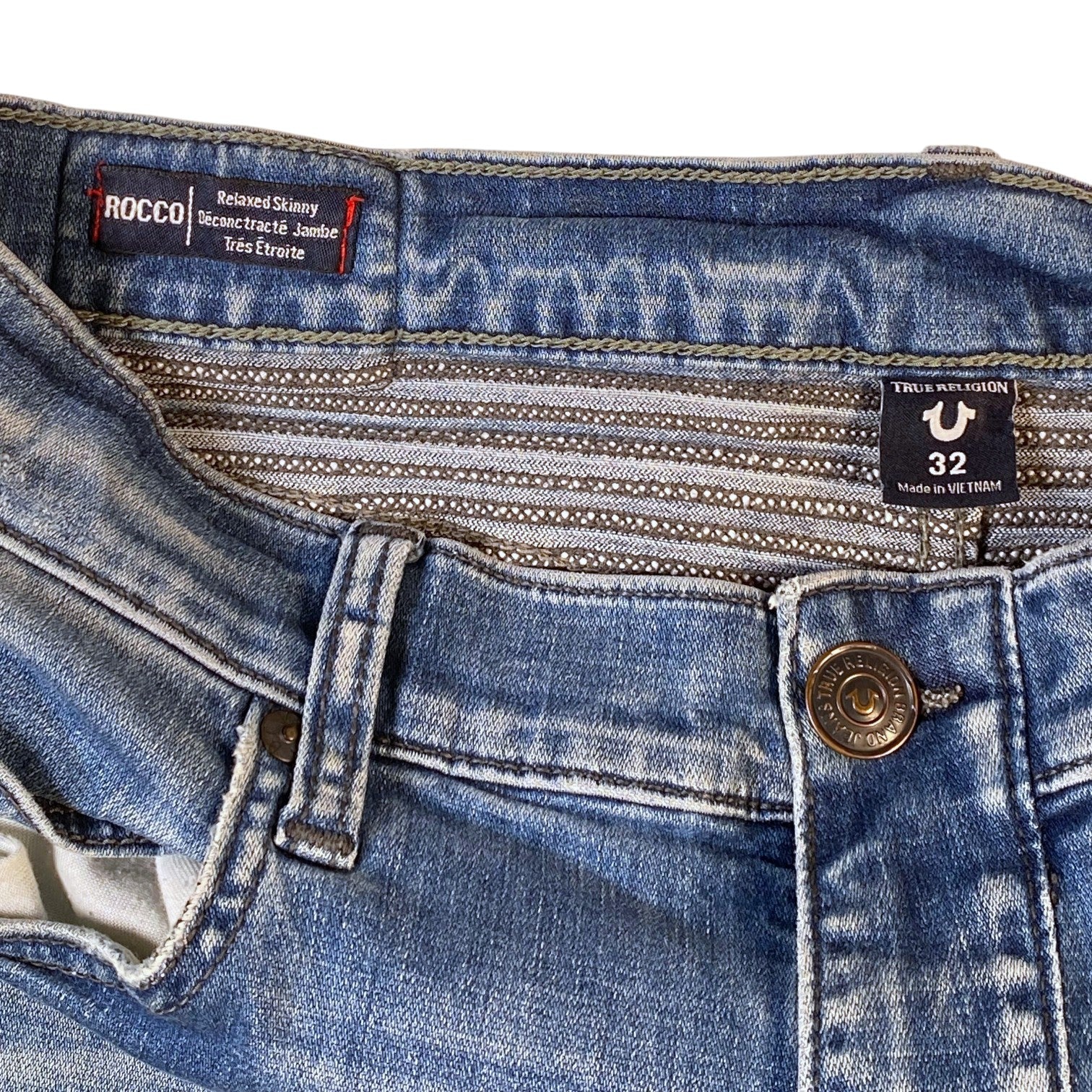 Close-up of the waistband and front closure of blue denim jeans, showing a metal button, belt loops, and two visible brand labels: Rocco and Trtl.