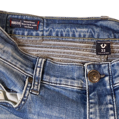 Close-up of the waistband and front closure of blue denim jeans, showing a metal button, belt loops, and two visible brand labels: Rocco and Trtl.