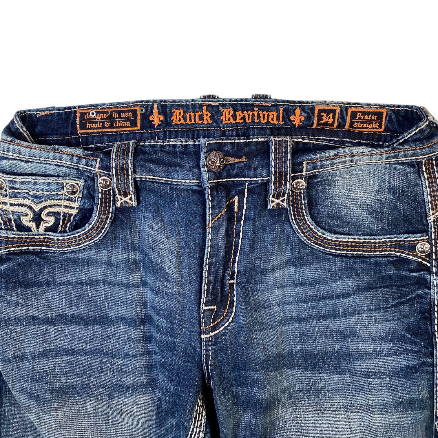 Close-up of blue denim jeans showing the waistband, front pockets, and decorative contrast stitching. The inner label reads 'Rock Revival' and '34'.
