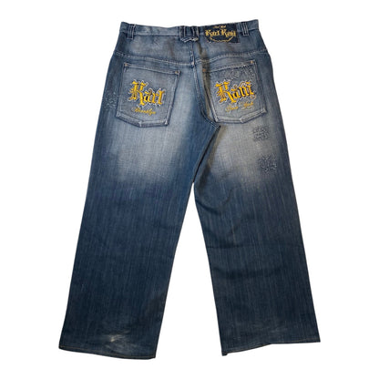 Blue denim jeans with a faded wash, featuring 'Korn' embroidered in gold on both back pockets and a matching 'Korn' label on the waistband, shown against a plain white background.