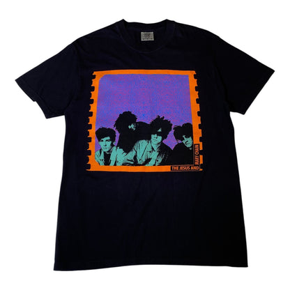 A black short-sleeve t-shirt featuring a vintage-style graphic print of the band The Jesus and Mary Children. The graphic has a purple background with a bright orange border and stylized greenish-blue portraits of the band members.