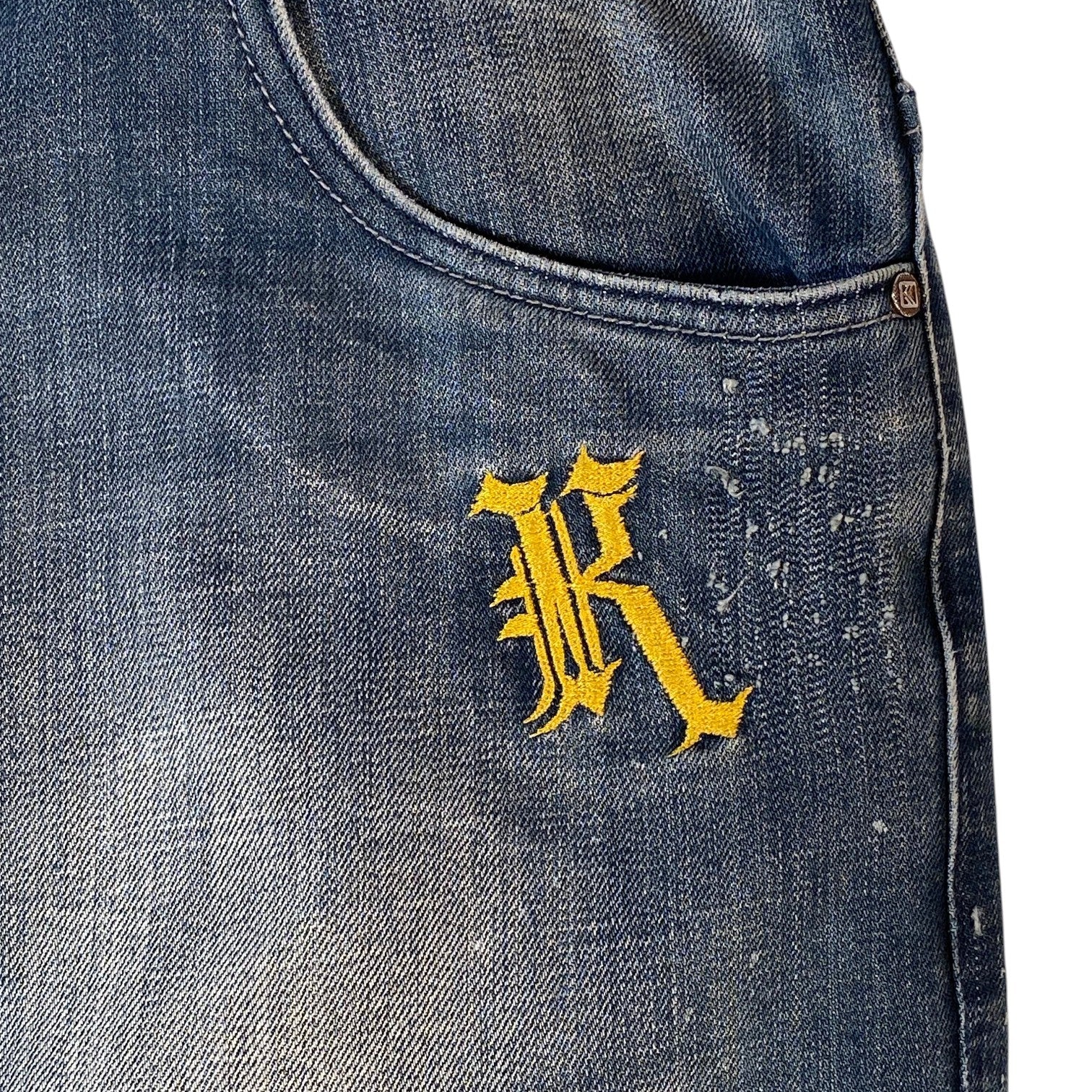 Close-up of a blue denim garment, likely a jacket or vest, featuring a yellow embroidered gothic letter 'R' on the chest pocket area.