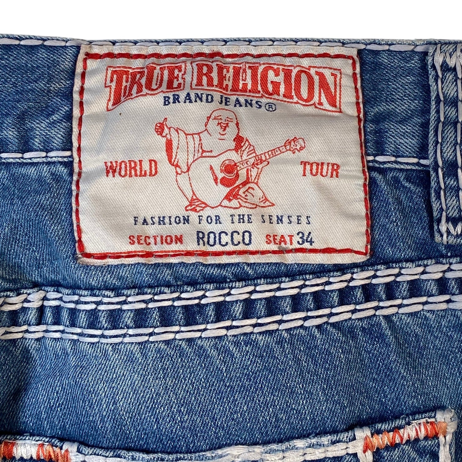 Close-up of the back waistband of a pair of True Religion Brand Jeans, showing the iconic red and white paper label with the Buddha graphic and 'World Tour' text, set against blue denim with white contrast stitching.