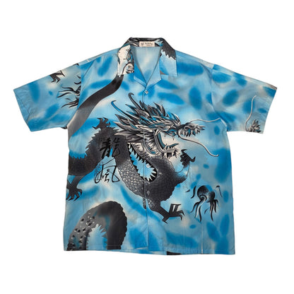 A short-sleeve button-up camp collar shirt featuring a large black and gray dragon illustration over a blue watercolor-style background, shown flat against a white background.